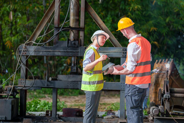 Male and female industrial engineers in Hard Hats discussing new projects while using laptops. They make gestures as they work in a heavy industrial factory.