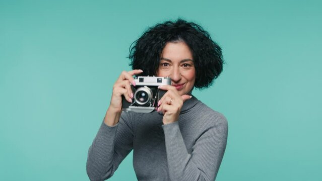 Portrait shot of smiling photographer with analog camera in teal blue photo studio on isolated background, looking at camera and viewfinder. Slow motion professional mature female photographer 4K