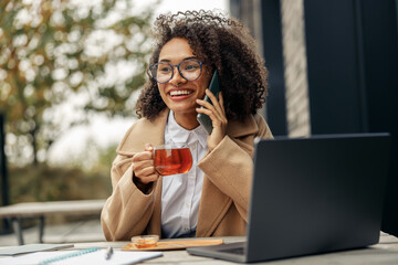 Smiling afro american businesswoman talking phone while working online sitting in cafe terrace