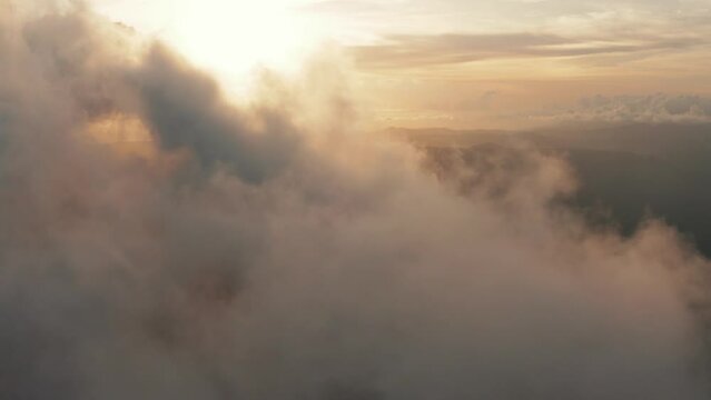 Aerial view flying through light, heaped up and wispy clouds in golden sunset, sunrise. Forward motion, slight pan to left. Underneath is mountainous landscape covered with forests. 4k footage.