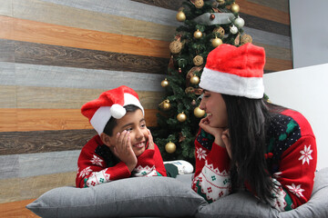 Single divorced mom and son Latino dressed in ugly sweters play in bed next to the Christmas tree with Santa hats give each other hugs and kisses show their love in solitude