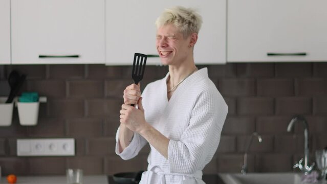 Young handsome happy man having fun in kitchen, listening to music and singing using kitchen spatula instead of microphone