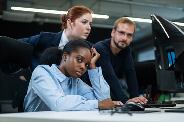 Obraz premium Colleagues look at the monitor and decide working moments. A sad African young woman is sitting at her desk, while a Caucasian man and a red-haired Caucasian woman are standing nearby.