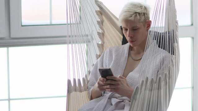 The guy in robe is sitting in a hammock on the balcony by window and looks down at the phone screen. He is resting and communicating