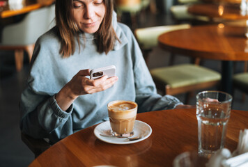 Young brunette woman wearing casual longsleeve with mobile phone in hands with cup of coffee sitting in cafe, city life