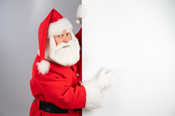 Santa Claus peeks out from behind a white background and points to an empty space with his finger. Merry Christmas. 