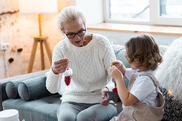 Grandmother and granddaughter doing craft toys and knitting near decorated Christmas new year tree. Cute little girl and attractive senior woman at home in the living room on the sofa having fun 