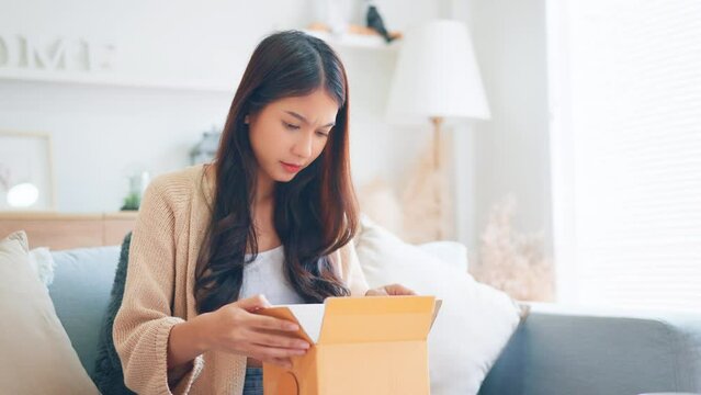 Wrong Item. Bad delivery service, Upset young asian woman unpacking cardboard box on sofa at home, receiving damaged item, Female feeling sad after unpacking box with delivery.