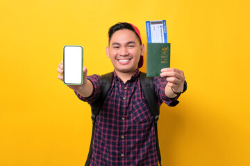 Cheerful young Asian man with backpack showing mobile phone blank screen, holding passport and flight tickets isolated over yellow background. Tourism and holiday trip concept