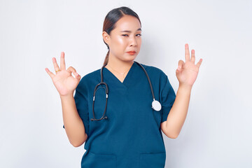 Smiling young Asian woman nurse wearing blue uniform with stethoscope, assures the quality of test results in their clinic lab, showing okay sign isolated on white background