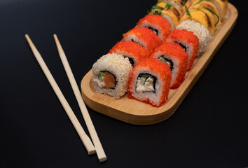 Delicious Japanese rolls with salmon, avocado, cucumber, cheese on wooden plate with wood sticks with selective focus on black background. Asian food concept. Healthy eating