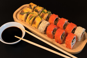 Delicious Japanese rolls with salmon, avocado, cucumber, cheese on wooden plate with wood sticks and soy sauce with selective focus on black background. Asian food concept. Healthy eating