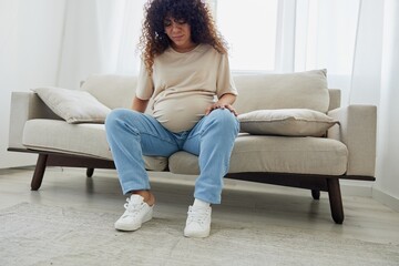 Pregnant woman sitting on sofa at home and putting on pregnancy-friendly shoes with problem due to belly
