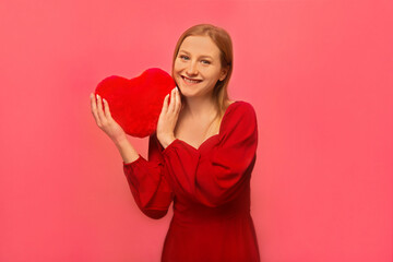 Happy smiling pretty blonde girl in red dress holding red heart toy and looking at camera isolated on colored pink background.

Happy Saint Valentines Day celebration concept.