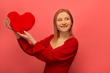 Happy smiling cheerful blonde girl holding red heart toy, wearing red dress with sleeves and looking up isolated on pink background.

Happy St Valentine's Day celebration concept.