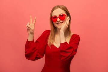 Stylish happy smiling young woman in red dress, french beret and trendy heart shaped glasses showing peace gesture v sign with two fingers and keeping hand on face isolated on colored pink background.