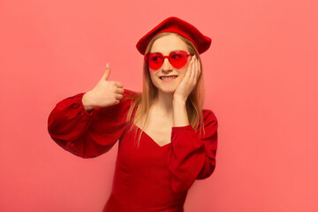Stylish happy smiling young woman in red dress, french beret and trendy heart glasses showing thumb up or holding finger up and keeping hand on face isolated on pink background.

St Valentines Day.