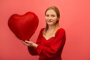 Cheerful stylish young woman holding heart shaped air balloon, wearing red dress with heart earrings and looking at camera on pink background.

Saint Valentines Day or birthday celebration concept.