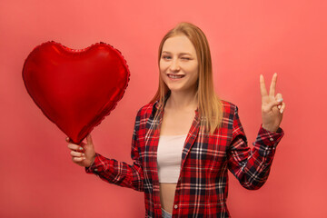 Cheerful happy winking girl showing peace gesture sign with two fingers and holding red heart...