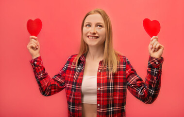 Happy smiling dreaming girl holding two red paper valentines heart cards, wearing checkered plaid shirt with white top and looking up isolated on pink background.

St Valentines Day or love concept.