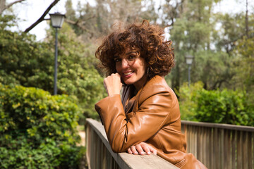 beautiful woman with curly hair leaning on the railing of a wooden bridge in a park with abundant vegetation The woman is dressed in modern and trendy clothes. Travel and holiday concept.