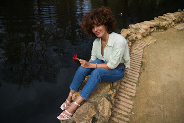 Beautiful woman with curly hair sitting on a duck and swan pond in the park. The woman is happy and holds a red flower in her hand as she looks at it excitedly.