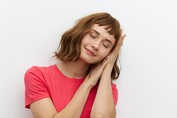 Obraz premium a gentle, relaxed, sleepy woman stands in a red T-shirt on a white background and holds her hands folded in her palms under her head, closing her eyes