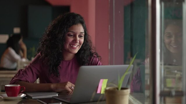 Young woman smiling and laughing while watching a funny video on laptop.Indian girl holding phone watching funny videos,movie or social media content while sitting in cafe.