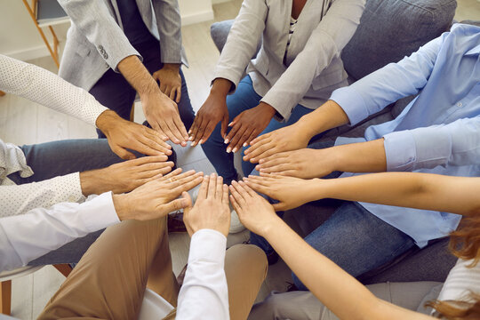 Different Multiracial Men And Women Form Circle With Their Hands Showing Their Unity And Equality. Cropped Image Of Hands Of People In Business Casual Clothes Sitting On Chairs In Circle.