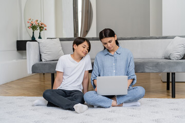 Asian boy sitting near mother working on laptop in living room