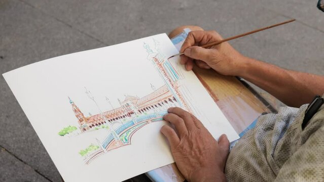Crop from above of senior male bearded artist in street drawing "Plaza de España, "Spain Square" of Seville, Spain building on paper with watercolor while sitting on chair