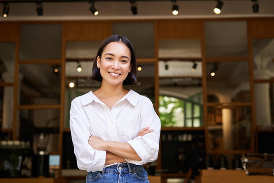 Smiling young female manager, cafe owner cross arms on chest, looking confident, standing in front of restaurant