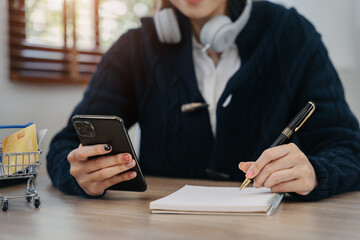 woman using smartphone to work and writing on notebook with pen in home office.