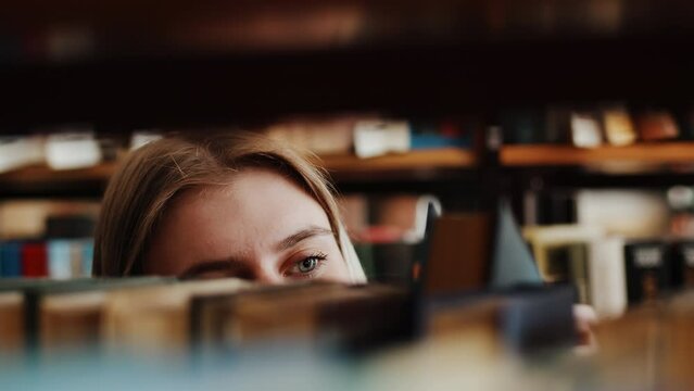 Young caucasian woman student standing between bookcases and thoroughly choosing a book used in her education from the shelf in the library close up