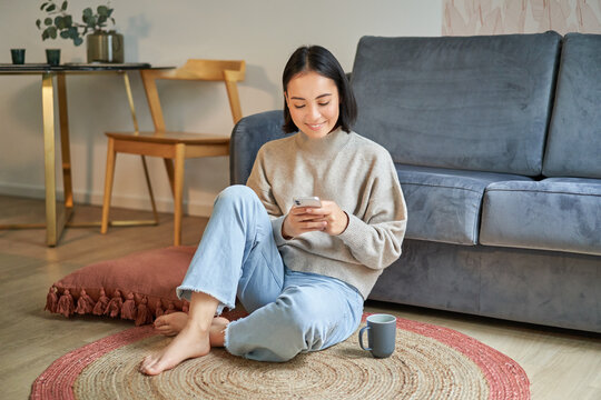 Image of stylish young woman in modern house, using mobile phone, sitting on floor and holding smartphone, drinking from cup