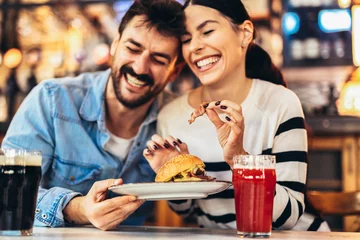 Foto op Plexiglas Young couple in love having fun spending leisure time together at restaurant, eating burgers and drinking beer © Mediteraneo
