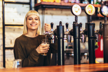 Female bartender tapping beer in bar. - obrazy, fototapety, plakaty