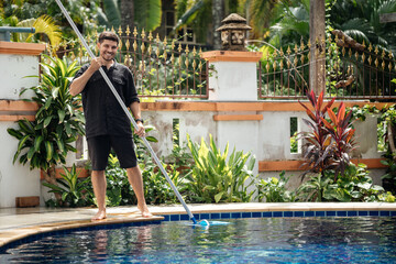 A pool cleaner in a black shirt and work shorts maintains the pool in the villa during the summer.