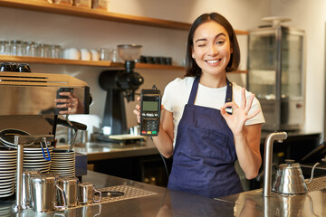 Smiling asian barista girl, wears apron, shows credit card machine for processing payment, suggest...