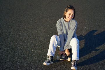 Portrait of beautiful asian girl skating, sitting on her skateboard and smiling. Cute teenager with...