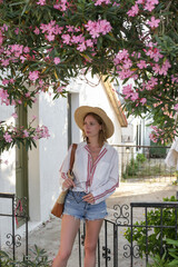 Young fashionable woman in cotton shirt and denim shorts with straw hat posing near blooming pink...