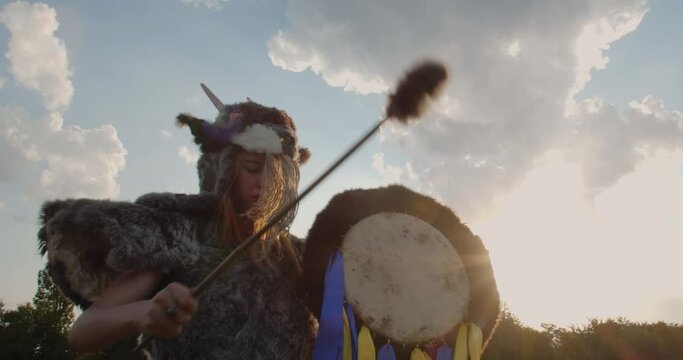 A blonde girl in a pagan fur costume made of animal skins and a goat mask with horns knocks on a leather tambourine against the background of an old wooden windmill and a rural landscape.