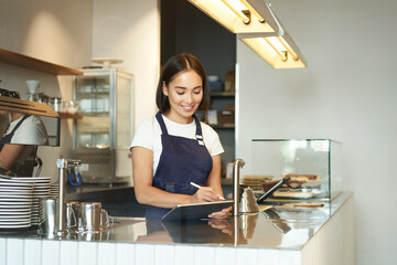 Portrait of beautiful asian girl smiling, barista in cafe working behind counter, using tablet as...
