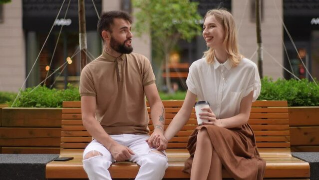 Portrait of happy young couple sitting on bench and drinking takeaway coffee enjoying time together outdoors. Pretty woman and bearded man having carefree talk sitting on bench holding hands.