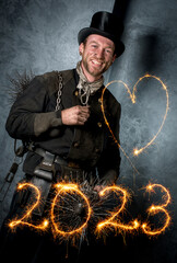Schornsteinfeger in Arbeitskleidung vor dunkler Wand, Jahreszahl 2023 und Herz mit strahlenden Wunderkerzen geschrieben.