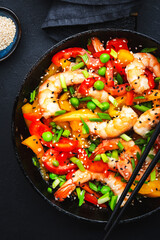 Stir fry with shrimps, red and yellow paprika, green pea, chives and sesame seeds in frying pan. Asian cuisine dish. Black stone kitchen table background, top view