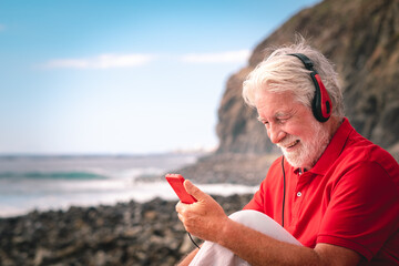 Beautiful senior man in red listening music by headphones sitting on the beach - elderly happy man...