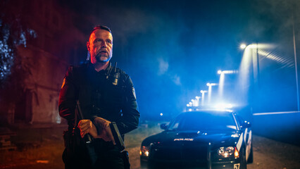 Portrait of Professional Caucasian Male Policeman Holding Flashlight and a Gun With a Police Car in...