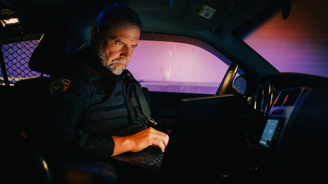 Inside Police Traffic Patrol Squad Car: White Male Police Officer On Duty Uses Laptop To Check Crime Suspect Background, License Plate, License And Registration. Officer Of The Law Fight Crime