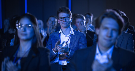 Crowd of Smart Tech People Applauding in a Dark Conference Hall During a Motivational Keynote Presentation. Business Technology Summit Auditorium Room Full of Delegates. Male in Focus.